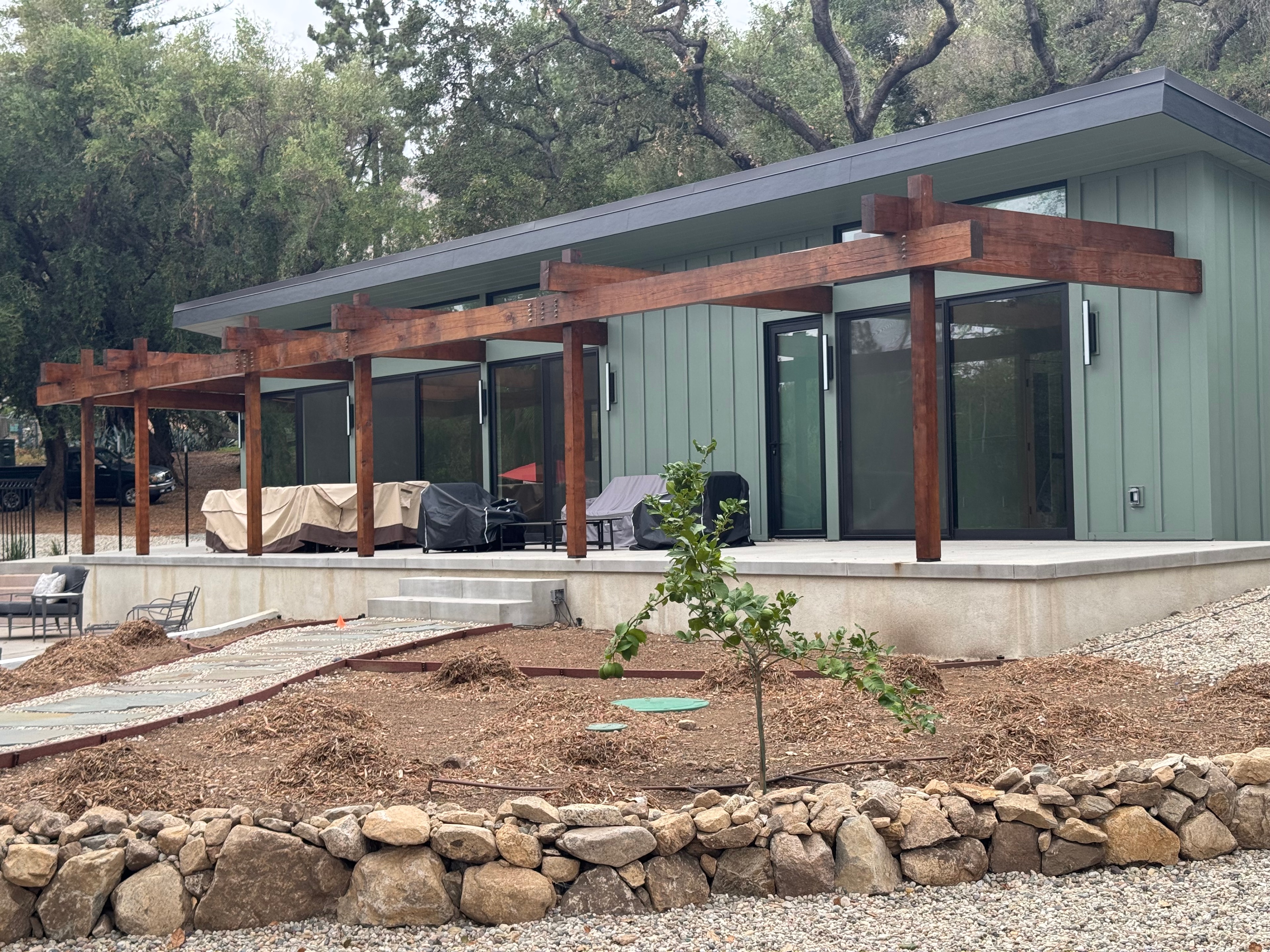 Completed modern ADU with concrete slab patio, wood pergola, and green board-and-batten siding, Los Angeles County